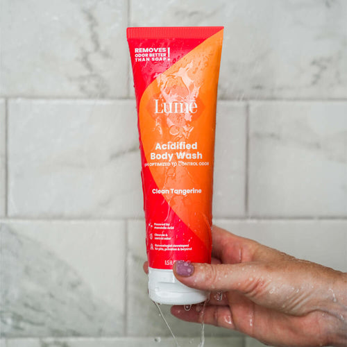 Hand holding a Lume clean tangerine scented acidified body wash inside a marble tile shower