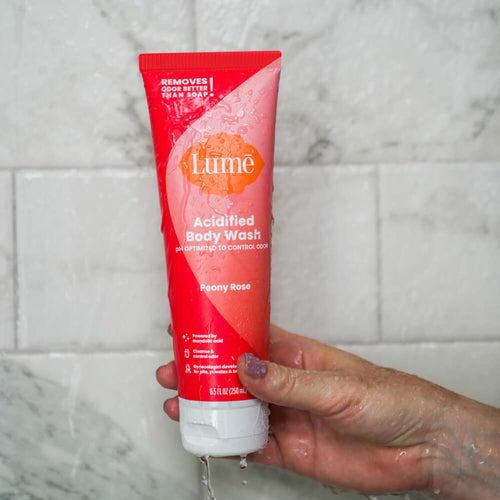 Hand holding a pink Lume peony rose scented acidified body wash inside a marble tile shower