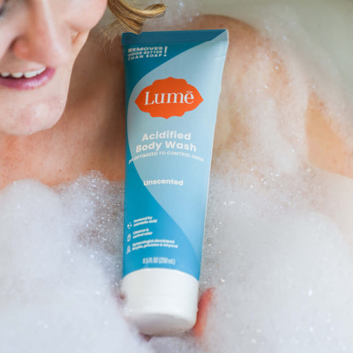 Woman taking a bath sorrounded by foam and holding a light blue Lume unscented acidified body wash tube