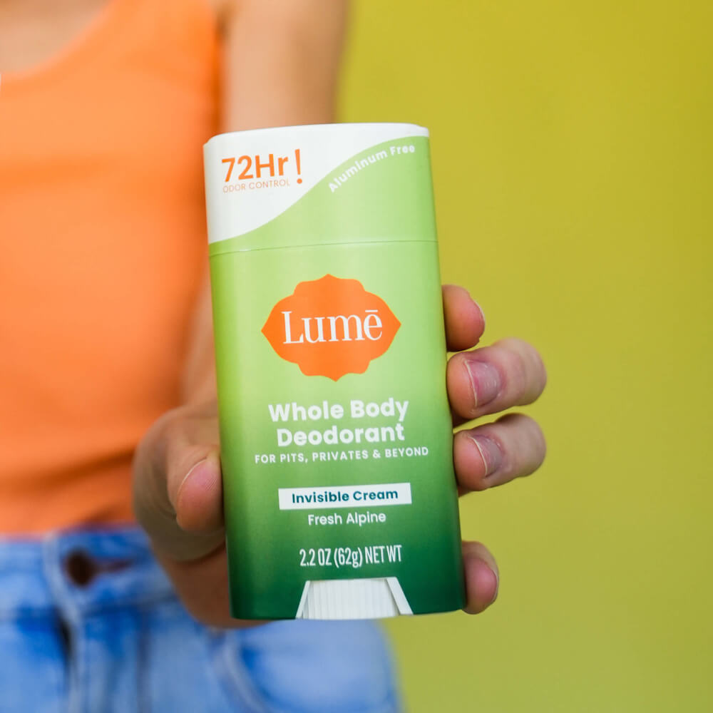 Woman in orange shirt and blue shorts holding up a green Lume fresh alpine scented cream deodorant stick