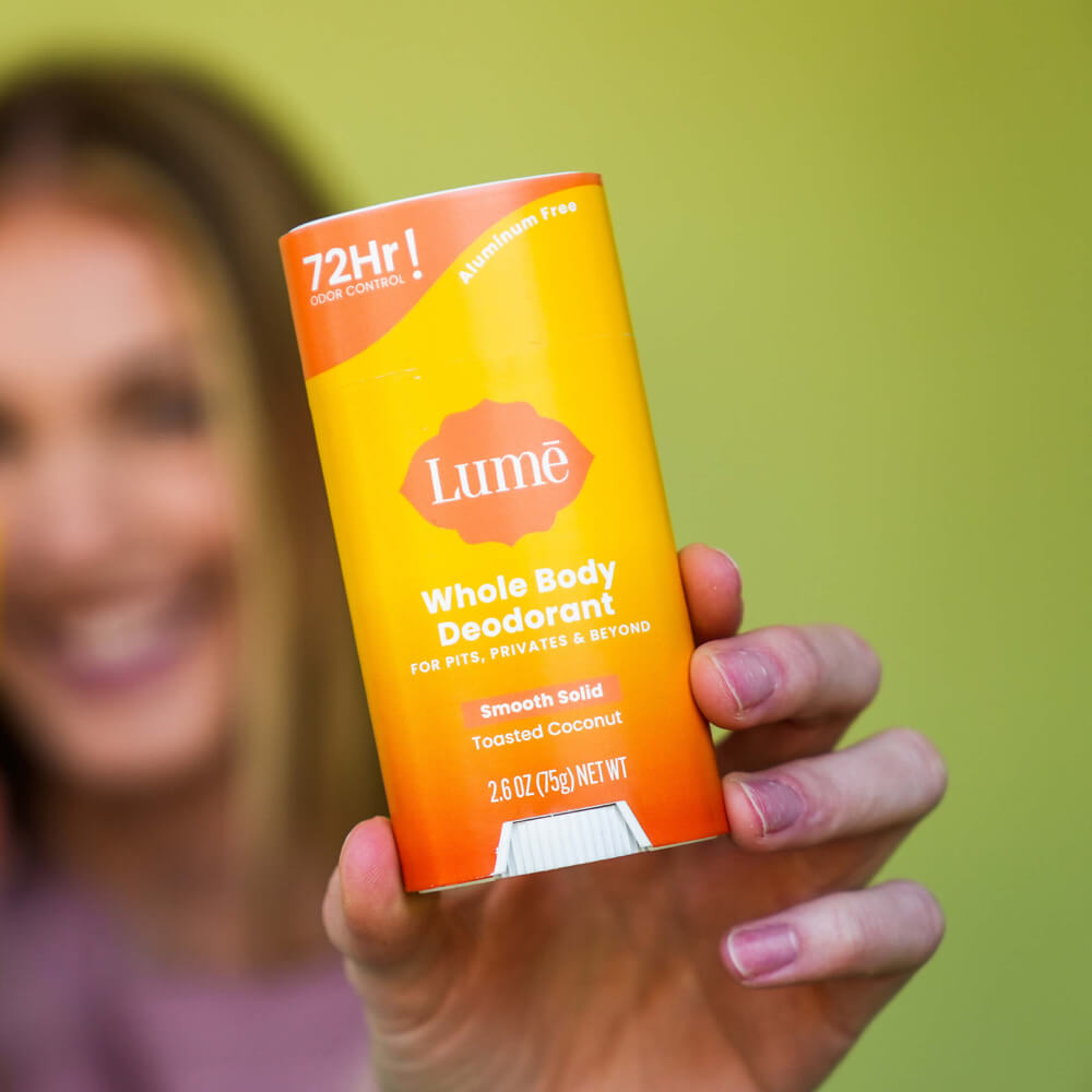 Woman holding up a bright orange Lume toasted coconut scented solid deodorant stick