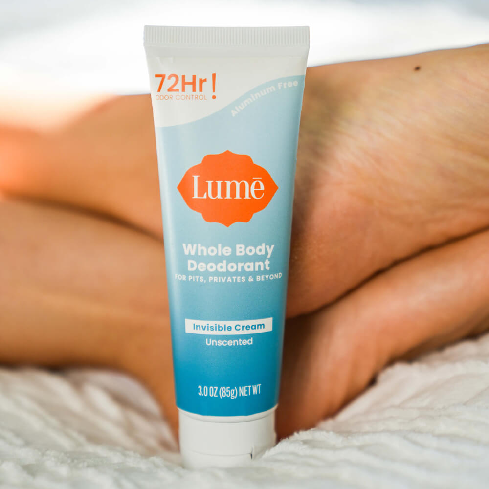 Blue and white Lume unscented cream deodorant tube on a white comforter next to a person&#39;s feet